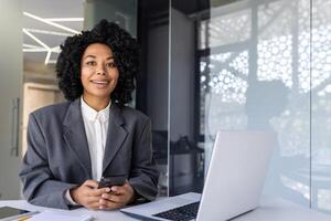 retrato de un joven exitoso afroamericano mujer de negocios sentado a un escritorio en un oficina a un escritorio, con confianza mirando a el cámara, participación un teléfono en su manos. foto