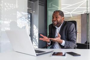 Angry nervous businessman talking remotely on call using laptop, mature experienced boss shouting and arguing sitting at workplace inside office. photo