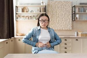 Young woman sick alone at home having severe stomach pain, asian woman holding her stomach with hands sad sitting on sofa in living room photo