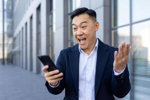 contento asiático empresario en pie fuera de en un traje y sorprendido y contento mirando a el pantalla de su móvil teléfono. foto