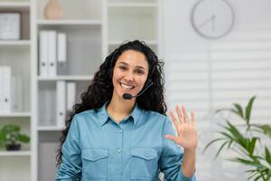 un alegre mujer en un azul camisa y auriculares olas a el cámara, retratar un acogedor en línea cliente Servicio ambiente en un moderno oficina. foto