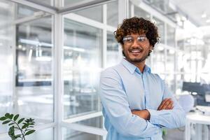 Portrait of young successful businessman inside modern office, hispanic man satisfied smiling and thinking, crossed arms and blue shirt and glasses looking window, good financier achievement. photo