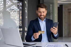 Smiling young businessman sitting in office at desk, holding phone and credit card, conducting online money transactions, transferring funds, checking account. photo