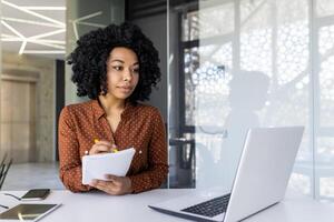 un enfocado joven africano americano mujer trabajando diligentemente en un brillante, moderno oficina espacio, tomando notas mientras consultante su ordenador portátil. foto