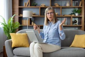 Mature woman at home looking at credit card with confusion, sitting on couch with her laptop, in a modern living room setting. photo