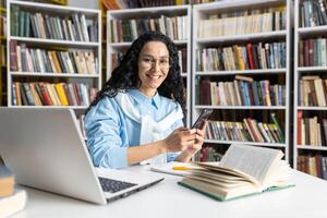 un alegre mujer vistiendo lentes interactúa con su teléfono inteligente mientras sentado en un biblioteca rodeado por libros. ella exuda un sentido de felicidad y contenido en un académico ajuste. foto