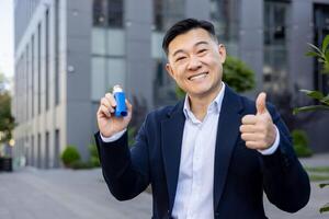 Portrait of a smiling young Asian man in a business suit sitting on a bench near an office building, holding an inhaler in his hand and showing the super sign with his finger to the camera. photo