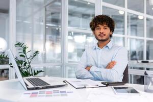 retrato de grave y confidente financiero empresario con cruzado brazos pensando mirando a cámara, exitoso Hispano hombre trabajando dentro financiero empresa oficina con documentos, utilizando calculadora foto