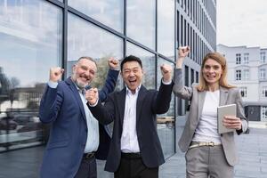 Successful diverse business team, three loving men and woman looking at camera and happy celebrating victory, team dreaming outside office building, business group in business suits photo