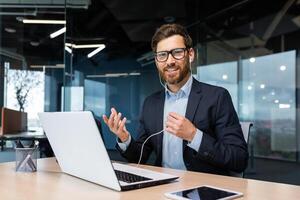 un joven empresario se sienta en el oficina a el mesa con auriculares, conduce un negocio reunión, un conferencia mediante un llamada desde un ordenador portátil. él olas su manos, mira a el cámara, sonrisas foto