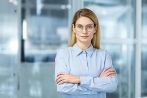 Serious ceo of financial company standing with arms crossed on chest and feeling proud about job success. Millennial female waiting for formal business meeting with investors in boarding room. photo