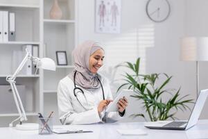 A cheerful female doctor wearing a hijab sits at her office desk, interacting with a digital tablet. The modern, bright workspace enhances her professional demeanor. photo