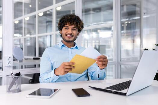 Portrait of a happy Hindu programmer inside the office, a businessman holds an envelope with a good news message in his hands, the man smiles and looks at the camera, uses a laptop at work. photo