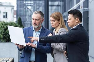 Successful business team, three colleagues businessman and businesswoman outside office building discussing current plans and management, looking at laptop screen, discussing and consulting photo