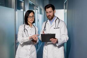 Two professional doctors in white lab coats looking at clipboard in hands while standing in hall of clinic. Coworkers discussing medical history of patient and forming new treatment strategy. photo