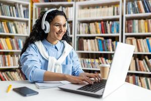 Smiling woman with headphones studying at a laptop in a library setting, surrounded by books and a coffee cup. photo