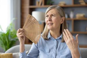 Disturbed woman waving with wooden hand fan to squinted face while sitting in overheated apartment. Exhausted woman suffering from no air conditioner at home during summer temperature increasing. photo