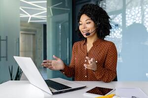 Young beautiful woman talking to customers remotely, african american female worker smiling using headset phone and laptop for online consultation, customer service. photo