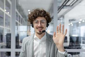 Smiling young businessman with curly hair and headset waving in office, representing a friendly corporate environment. photo