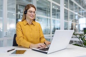 sonriente mujer de negocios trabajando dentro oficina con ordenador portátil y auriculares para llamar, mujer sentado a lugar de trabajo contento trabajando con clientela foto