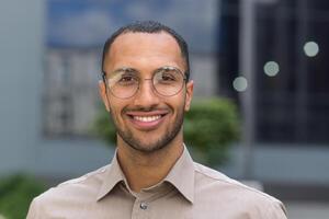 Close-up photo portrait of young entrepreneur wearing glasses, hispanic man smiling and looking at camera, startup entrepreneur outside modern office building