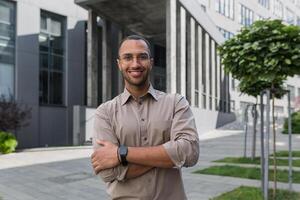 Young successful businessman smiling and looking at camera with crossed arms, man outside office building wearing shirt and glasses, startup investor photo