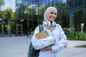 un joven estudiante en un hiyab, auriculares alrededor cuello, participación libros, en pie con orgullo en frente de un instalaciones edificio. foto