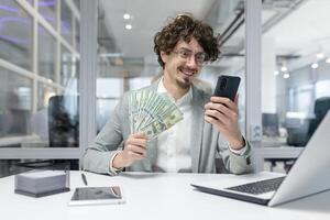 alegre joven profesional con Rizado pelo hace alarde ganancias mientras atractivo con un teléfono inteligente en un moderno oficina ajuste. foto