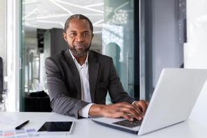 retrato de grave pensando y confidente empresario, africano americano jefe pensando y mirando a cámara, hombre trabajando dentro oficina en negocio traje, inversor financiero en con ordenador portátil. foto