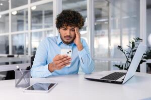 empresario dentro el oficina a el lugar de trabajo es no satisfecho con el trabajo de el teléfono, el hombre es trastornado participación un teléfono inteligente en su manos, Hispano hombre es sentado a el mesa con un ordenador portátil. foto