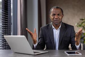 Perplexed African American businessman in a suit gestures with uncertainty at his office desk with laptop and tablet. photo