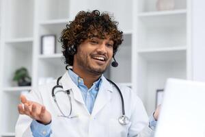 Young Indian doctor consulting patients remotely, man in white medical coat with headset using laptop for call, smiling contentedly inside clinic office. photo