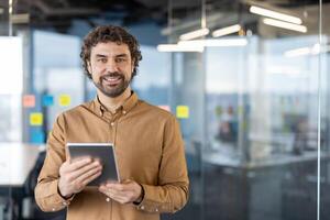 Portrait of a mature Hispanic businessman smiling confidently while holding a digital tablet in an office setting with glass walls. photo