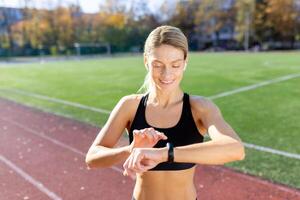 Female athlete smiling and checking results of fitness training and running on smartwatch bracelet, woman athlete smiling checking heart rate on device while running in stadium on sunny day. photo