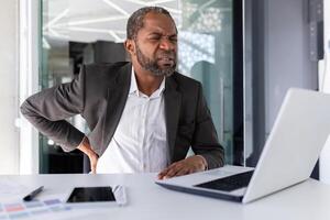 enfermo maduro africano americano empresario teniendo grave espalda dolor, hombre en negocio traje a lugar de trabajo sentado trabajo sobrecargado de trabajo y cansado. foto