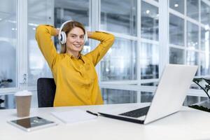 retrato de contento hembra programador dentro oficina, hembra trabajador con ordenador portátil y auriculares sonriente y mirando a cámara, mujer de negocios satisfecho con trabajo y logro resultados a lugar de trabajo. foto