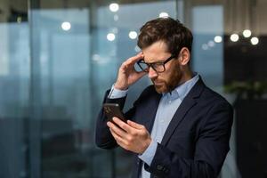 A worried young man businessman holds the phone in his hands, looks. Reading bad news, received a message. Shocked, holds his head. photo