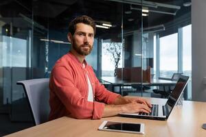 Portrait of mature programmer team leader man with beard and in red shirt is concentrating and thinking looking at camera , developer working inside modern company using laptop for coding. photo