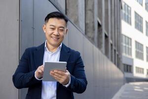 sonriente asiático empresario en un azul traje utilizando un digital tableta fuera de un oficina edificio en un soleado día. foto