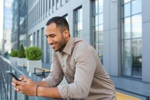 A young handsome Latin American man is standing on the veranda of the office center, holding a phone photo