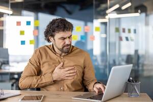 Adult male office worker with discomfort, clutching chest in pain while working on laptop, showing symptoms of stress or heart attack. photo