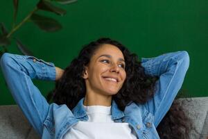 Beautiful hispanic woman daydreaming sitting on couch at home and looking out window, happy woman with hands behind head close up. photo