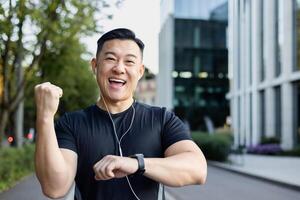Portrait of a positive and happy young Asian male sportsman who is happy about the result of a run showing a smart watch, showing a success sign with his hand, smiling at the camera. photo