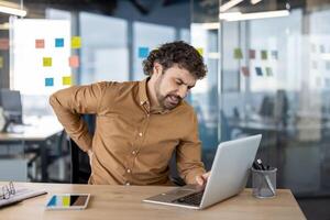 Adult male in office setting feeling discomfort in his lower back while sitting and working on his laptop, showing signs of pain. photo