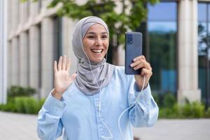 sonriente árabe mujer de negocios en hijab ondulación durante un llamar. joven hembra estudiante o oficina trabajador fuera de moderno edificio. foto
