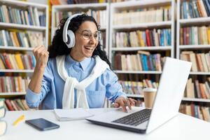 Joyful young student with headphones celebrating academic success in front of a laptop at the library, surrounded by books. photo