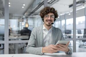 carismático joven profesional con Rizado pelo y auriculares con confianza trabajando en un contemporáneo oficina ambiente, utilizando un tableta. foto