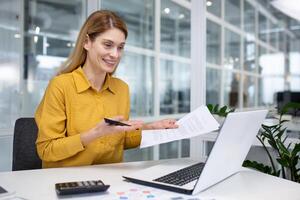 Mature business woman on online presentation, female financial worker on backup job shows documents reports to camera, uses laptop for call, remote meeting, works inside office. photo
