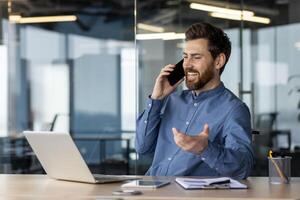 un profesional hombre visto mitad de la conversación en su teléfono inteligente, sonriente con un ordenador portátil y oficina suministros alrededor a él, ejemplificando un positivo negocio llamar. foto