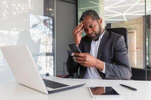 trastornado y decepcionado hombre dentro oficina participación teléfono, empresario leyendo malo Noticias a lugar de trabajo dentro oficina, africano americano mayor jefe infeliz con logro resultados notificación. foto